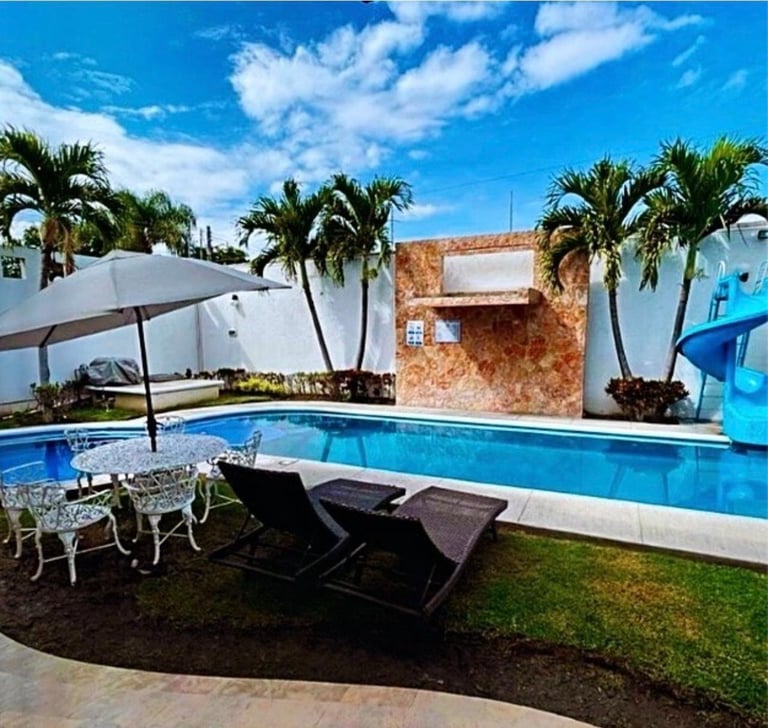 Modern resort backyard with rectangular swimming pool, black lounge chairs, white umbrella, palm trees, and rustic brick wall under blue sky