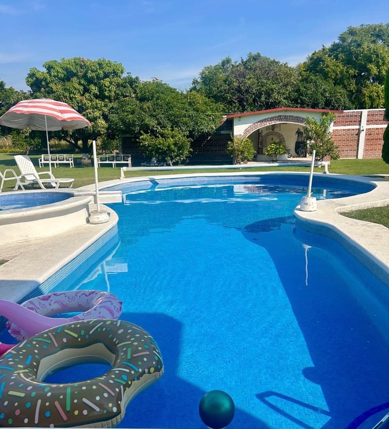 Bright blue swimming pool with floats, lounge chairs, umbrella, and tree-lined background on a sunny day