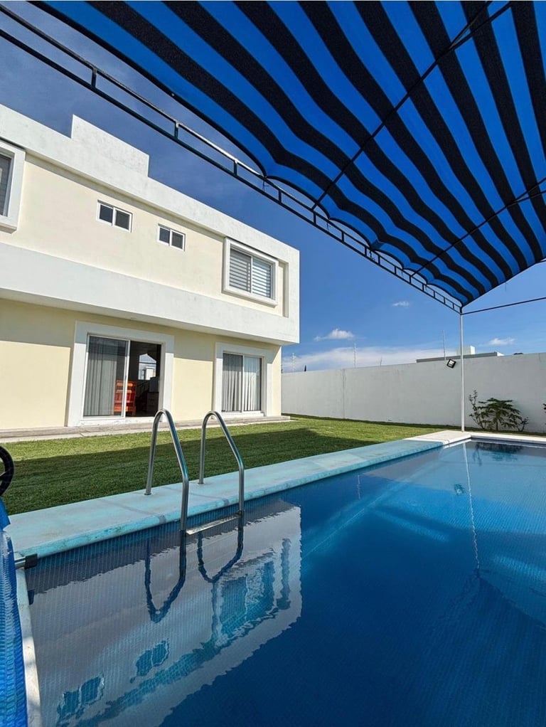 Modern house with blue and white striped awning overlooking a backyard swimming pool with metal ladder and manicured grass